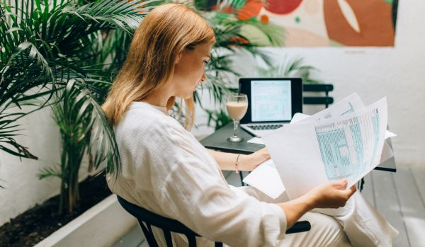 We are looking at the side profile of a woman reviewing a paper report. She is sitting next to a table with her laptop and a coffee.