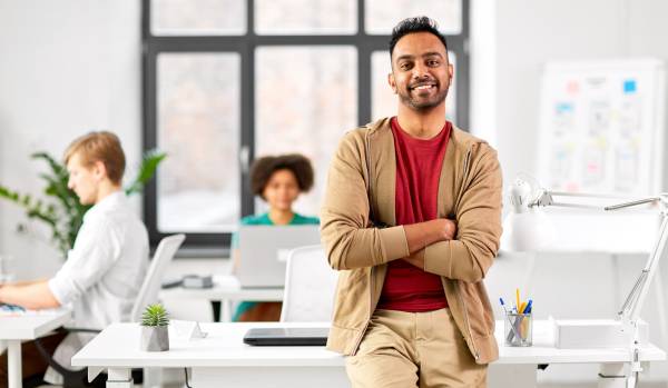A smiling young man in a casual outfit is standing with his arms crossed, leaning back on his desk. Behind him, two other people work on their laptops at their own desks.