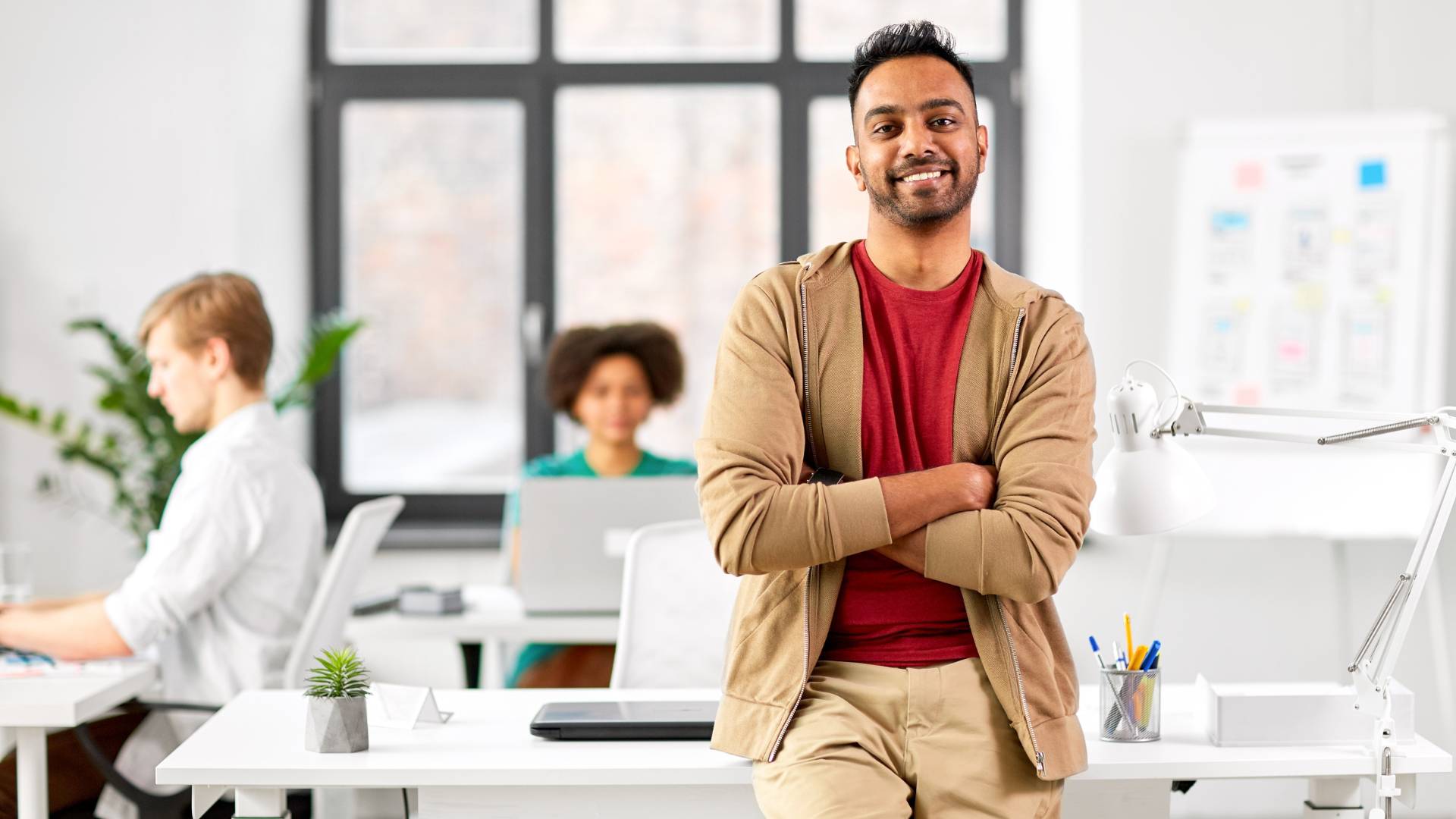 A smiling young man in a casual outfit is standing with his arms crossed, leaning back on his desk. Behind him, two other people work on their laptops at their own desks.