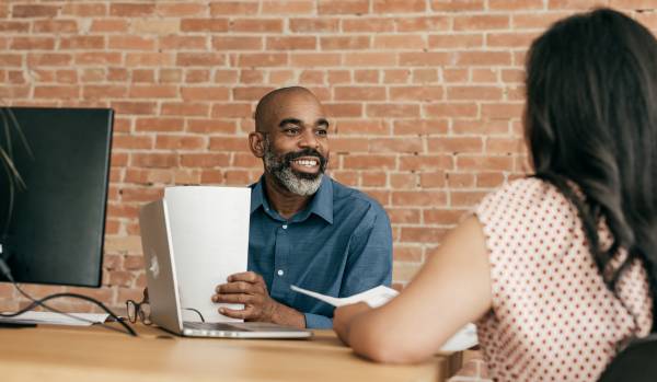 A Black man is sitting at a desk across from a woman with black hair. He is smiling while looking at her.