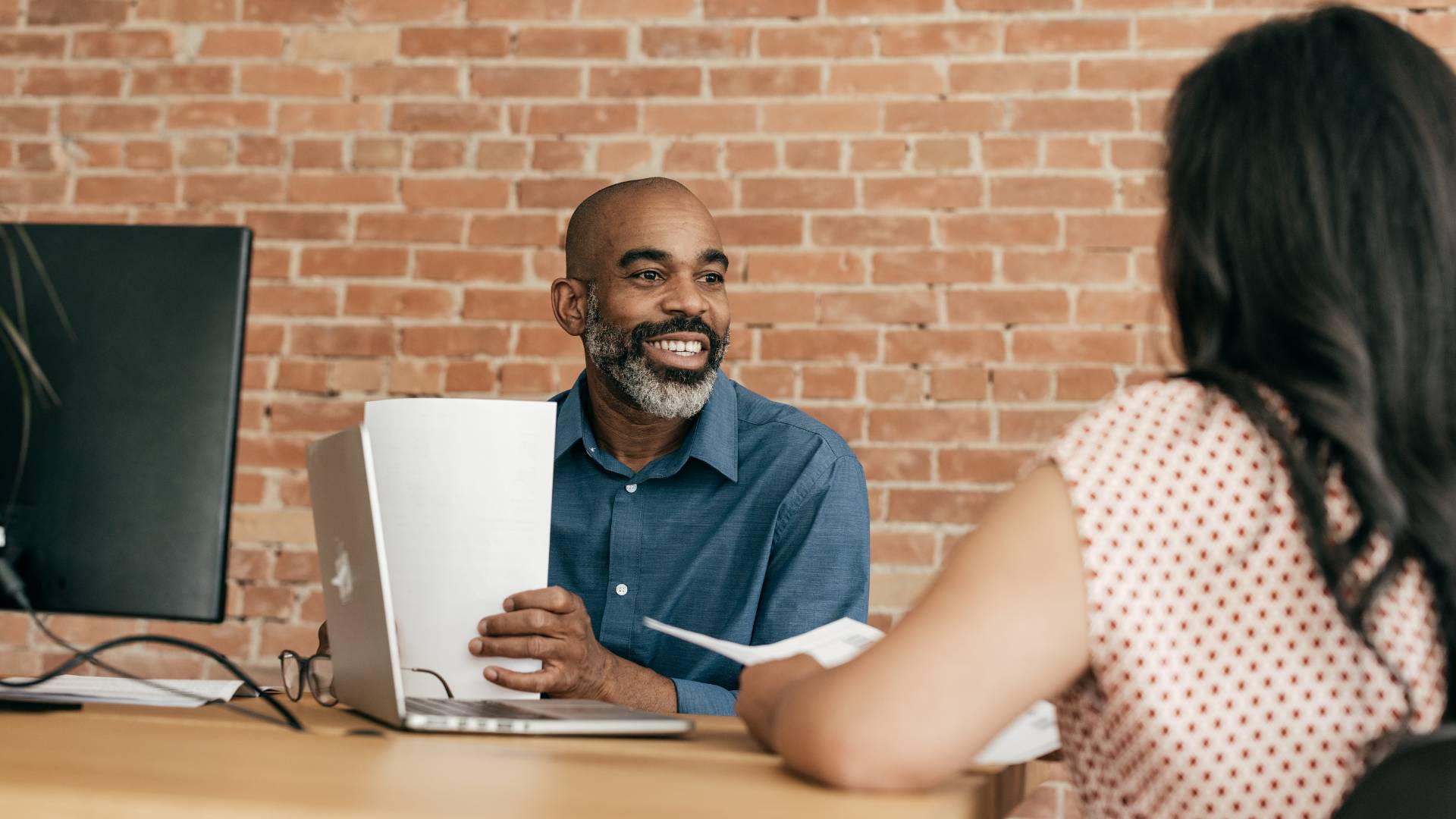 A Black man is sitting at a desk across from a woman with black hair. He is smiling while looking at her.