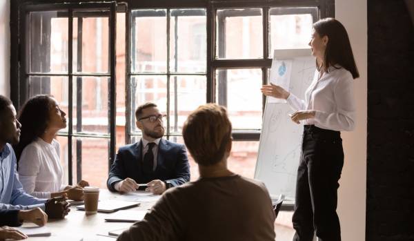 A woman who is a corporate trainer is leading a training inside of a conference room in an office setting. There are four people in the training.