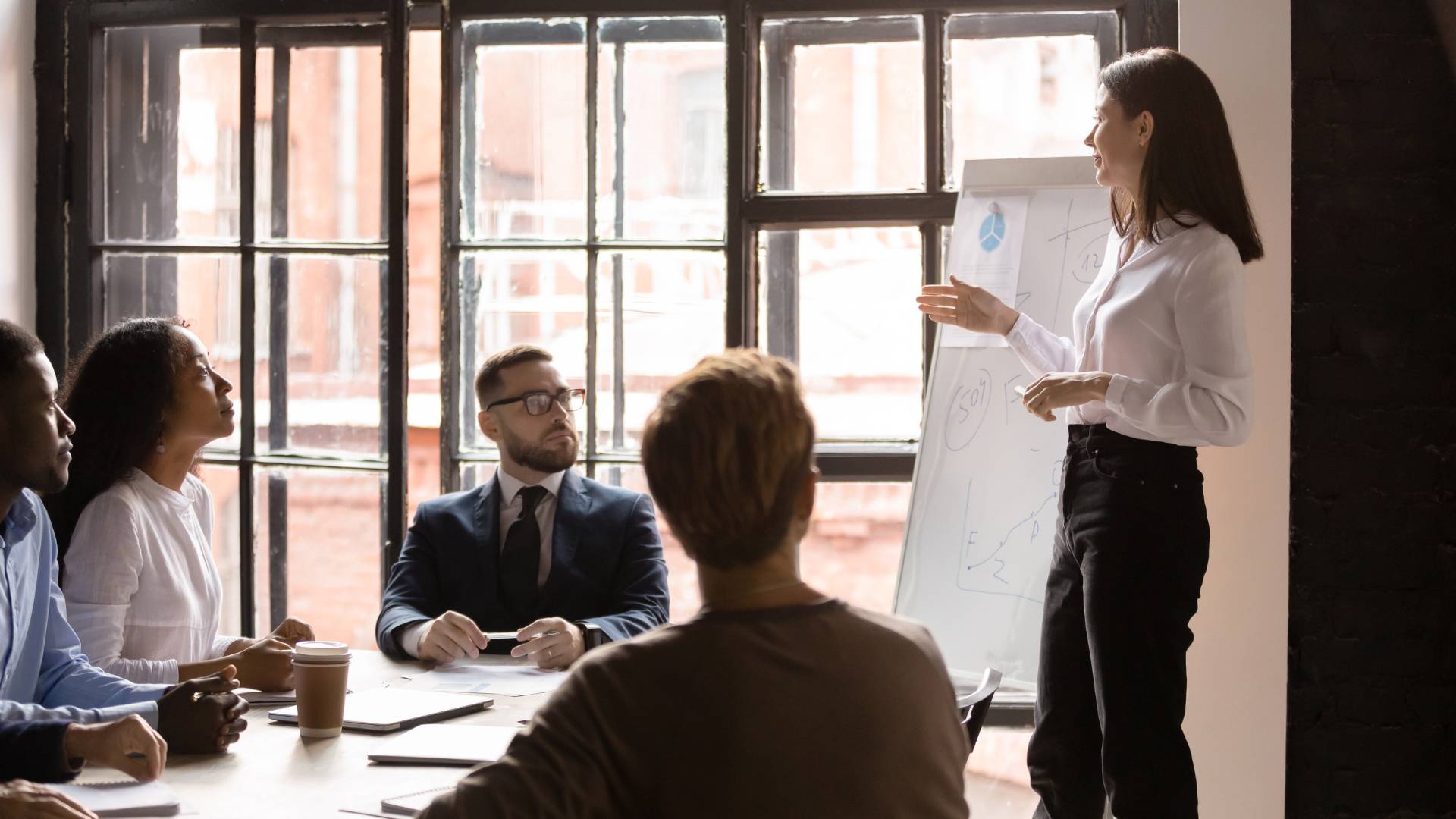 A woman who is a corporate trainer is leading a training inside of a conference room in an office setting. There are four people in the training.