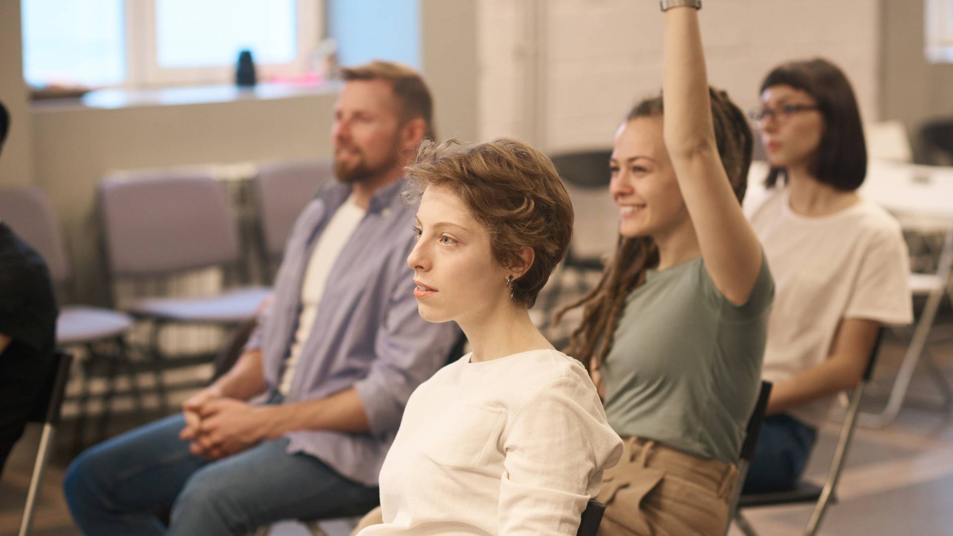 There are four employees sitting in a classroom setting in a training. One woman has her hand raised.