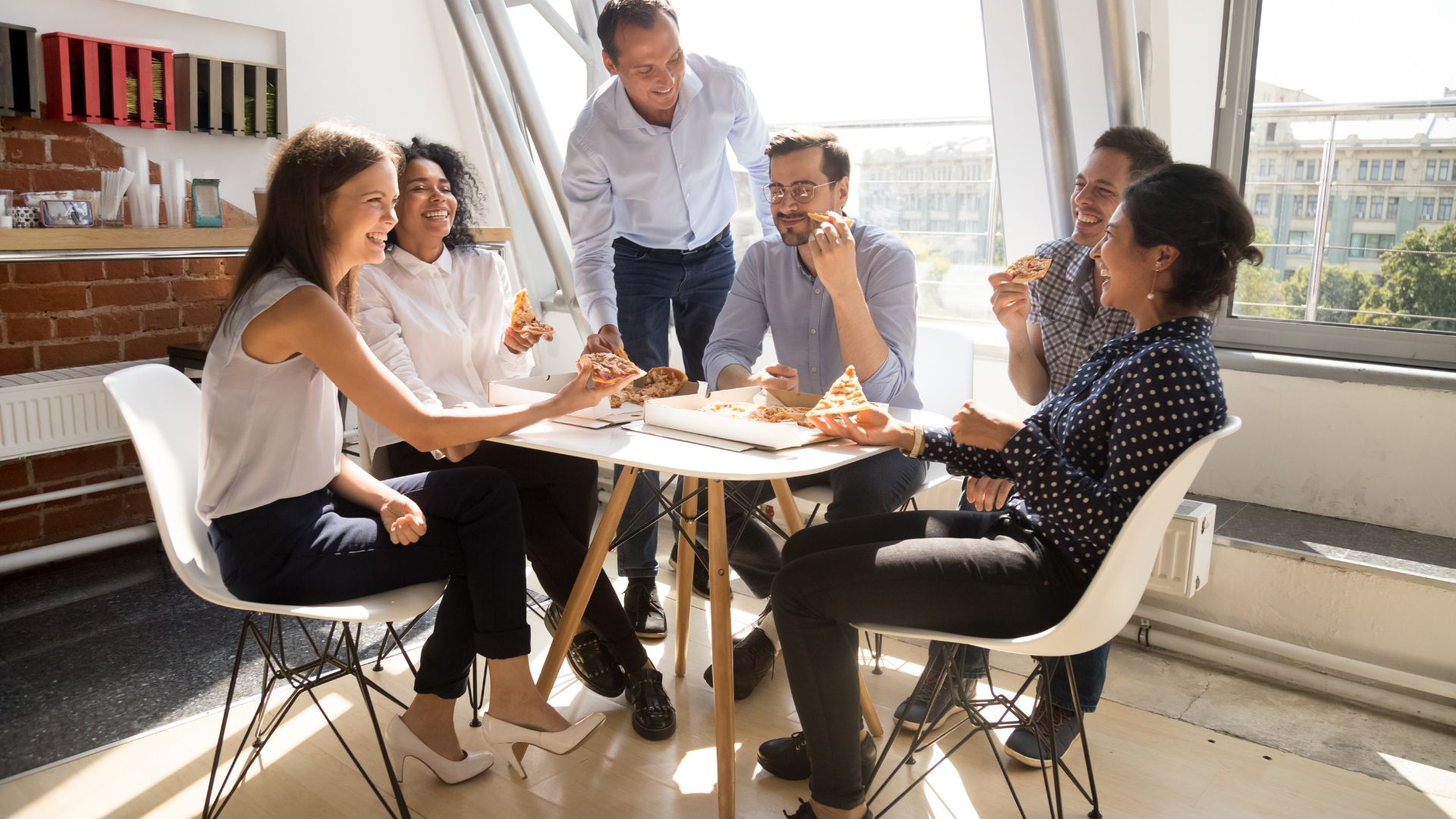 A diverse group of employees are laughing and smiling sitting and standing around a table in a common area, sharing a pizza.