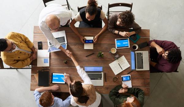 Overhead shot looking down at a table of diverse people working on their laptops, smiling and shaking hands while reviewing data.
