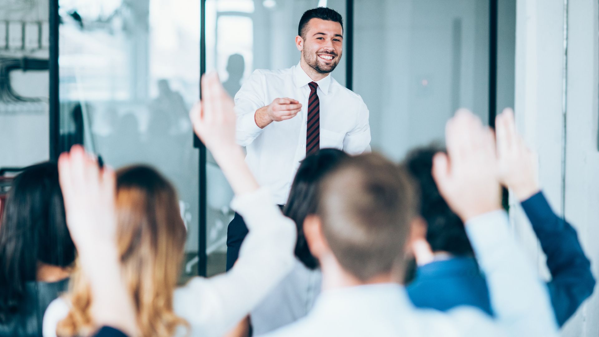 A man is standing in front of a room of professionals leading a training. We are looking over the participants with raised hands at the face of the man standing up front.