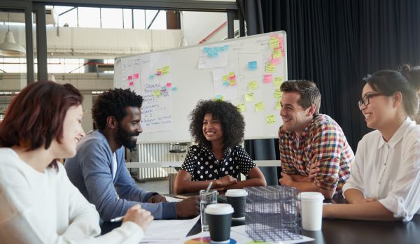 A diverse group of coworkers are smiling around a conference table with a whiteboard in the background.