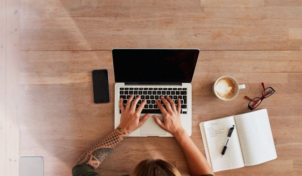 We're looking down from overhead of a person using their laptop. There is a notebook and coffee on the table next to them.