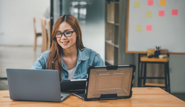 A young woman with glasses is smiling down at her two laptops during a remote professional development class.