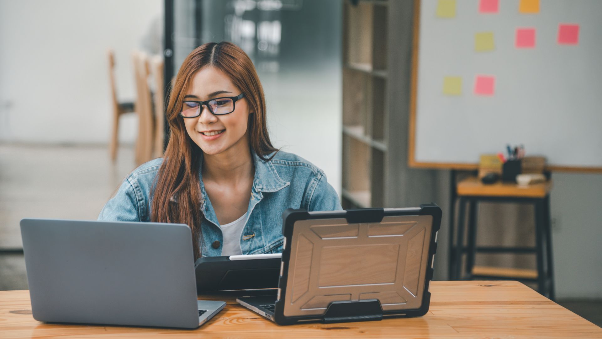 A young woman with glasses is smiling down at her two laptops during a remote professional development class.