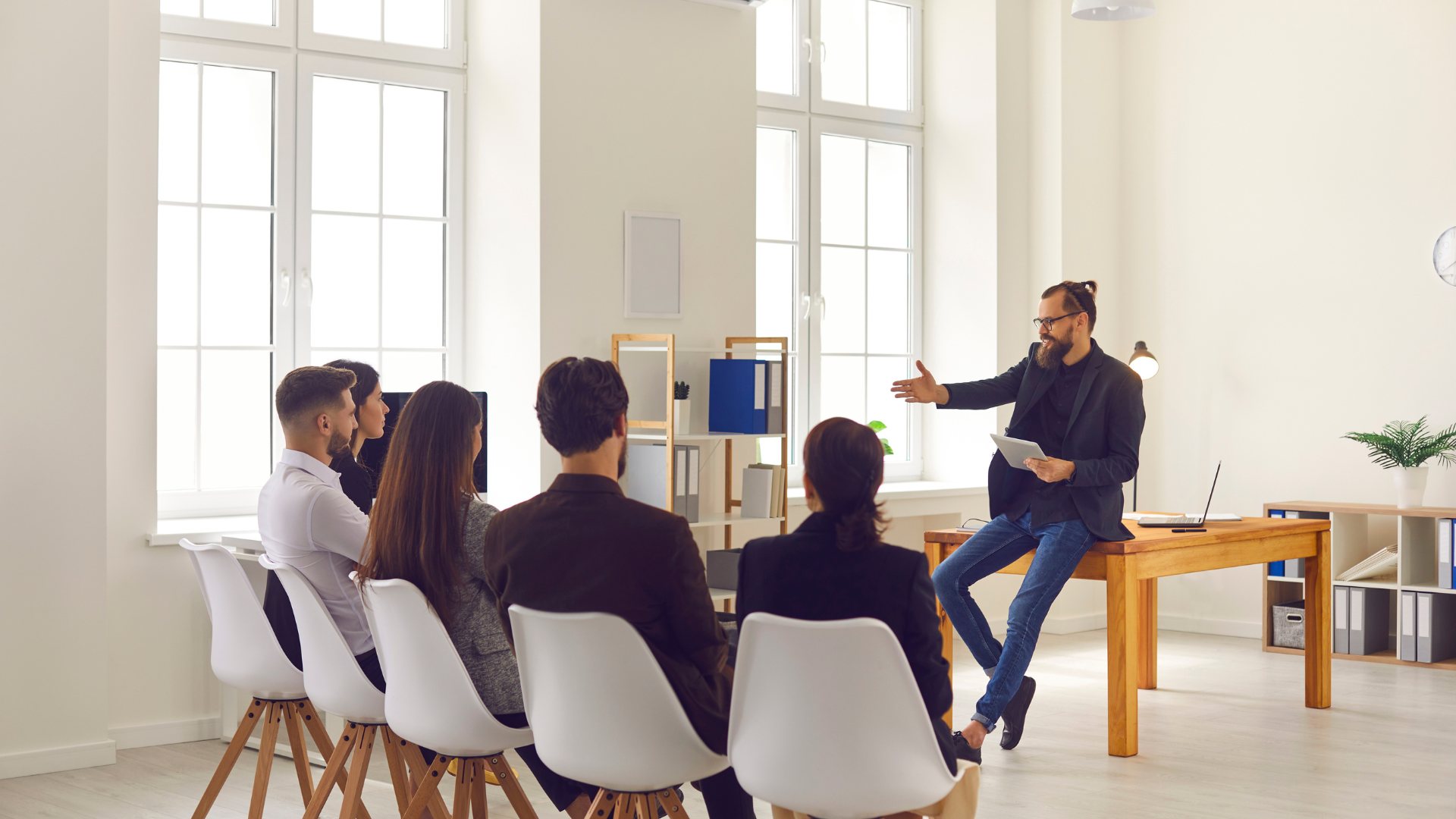 There are five people sitting in chairs in front of a man speaking who is leaning on a desk in an office setting.