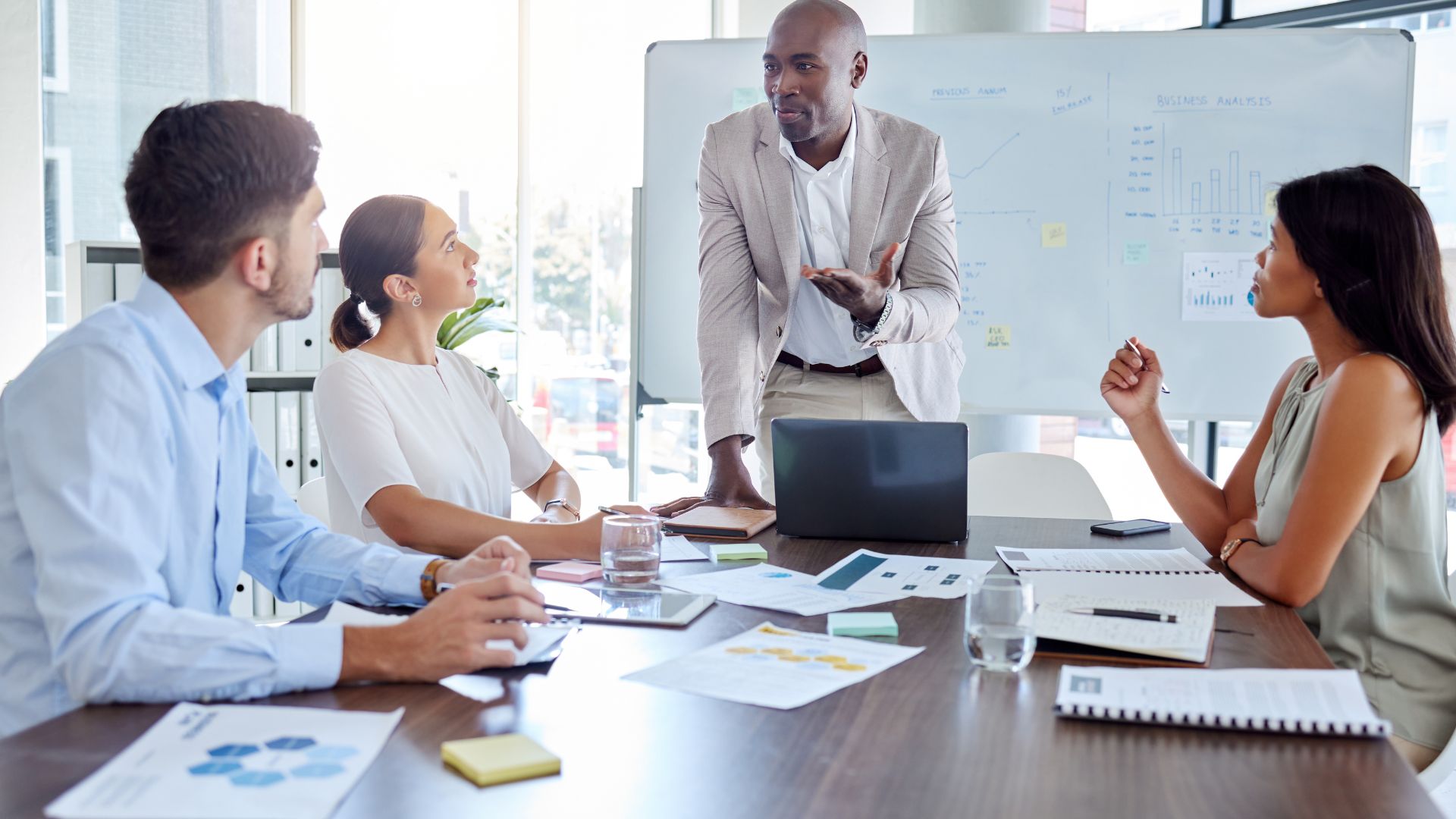 A man is standing at the head of a conference table teaching 3 other employees who are sitting around the table.