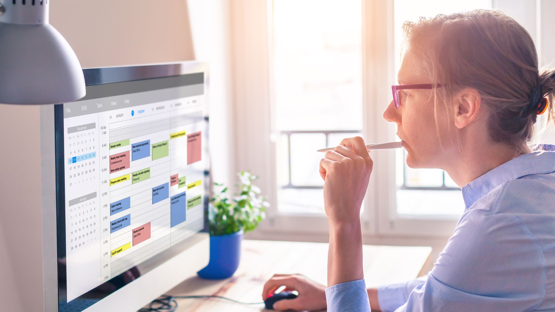 A woman in glasses is biting the end of her pen while looking at her calendar on her computer monitor.