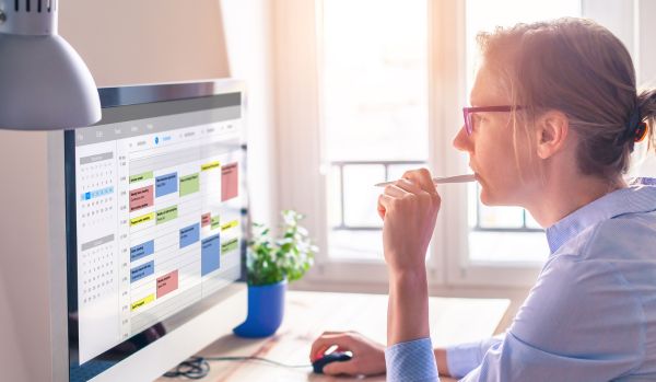 A woman in glasses is biting the end of her pen while looking at her calendar on her computer monitor.