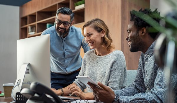 There are three people gathered around a computer looking at it and smiling. There are two men and one woman.