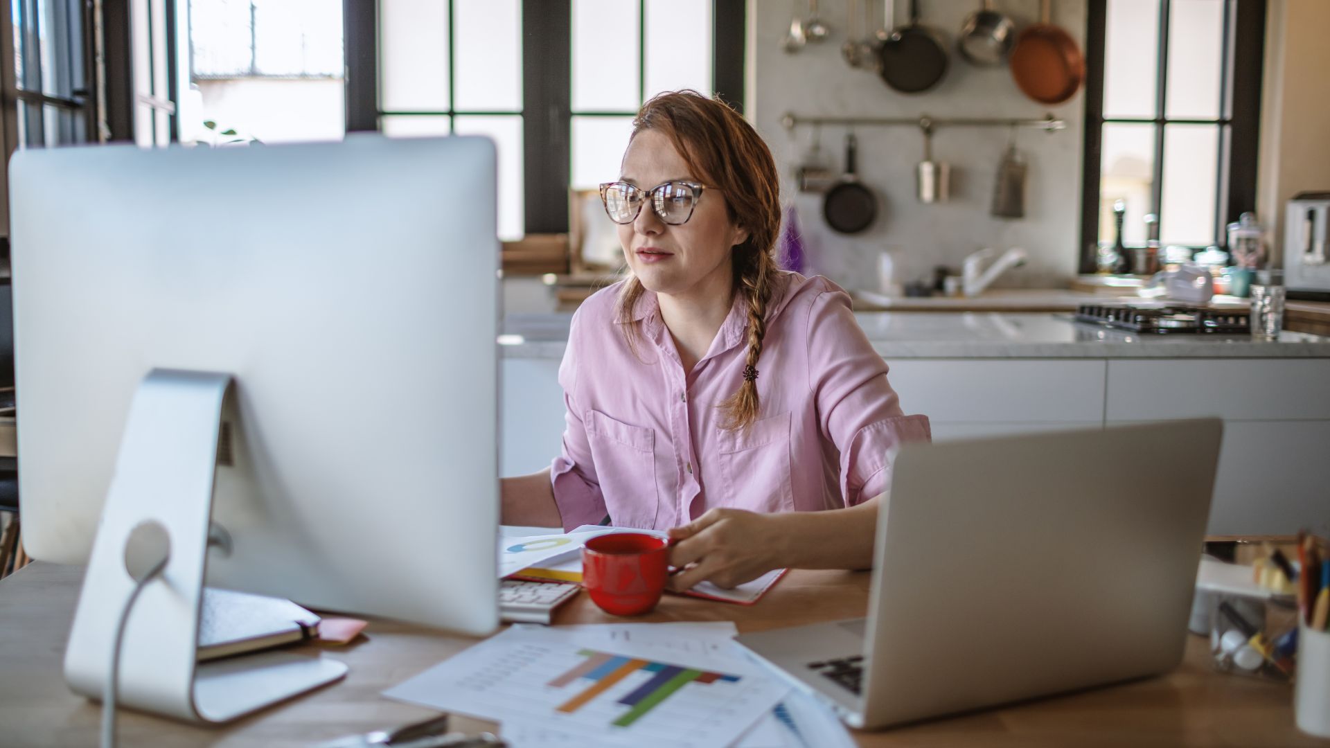 A woman is working on two computers from her kitchen table. She is holding a cup of coffee.