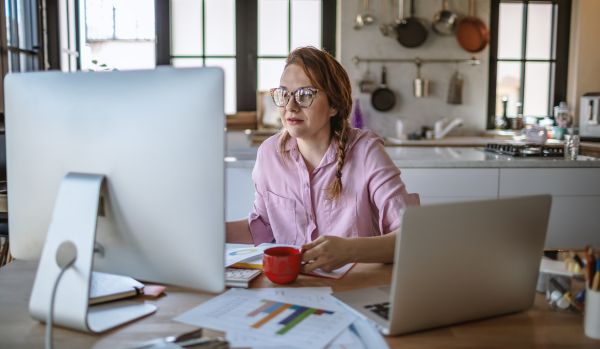 A woman is working on two computers from her kitchen table. She is holding a cup of coffee.
