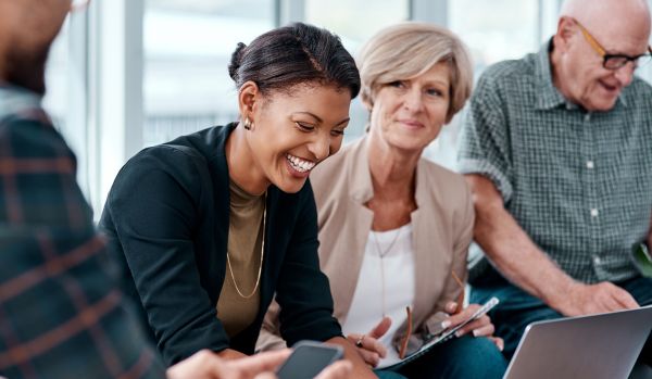 There are three people sitting next to each other in an office. There's a young Black woman laughing and sitting next to her is an elderly woman and an elderly man.