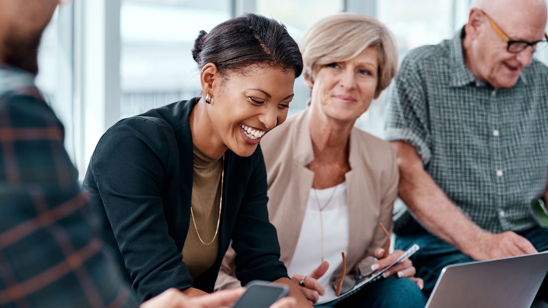 There are three people sitting next to each other in an office. There's a young Black woman laughing and sitting next to her is an elderly woman and an elderly man.