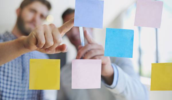 We're looking through a clear whiteboard at two people discussing a series of post-it notes as part of their design thinking process.