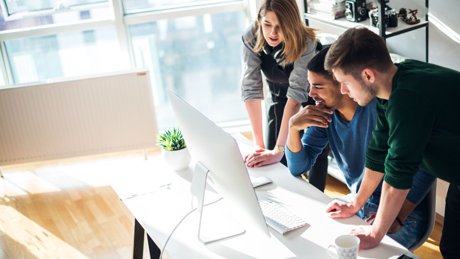A group of three people are gathered behind one person's desk looking at that person's computer monitor.