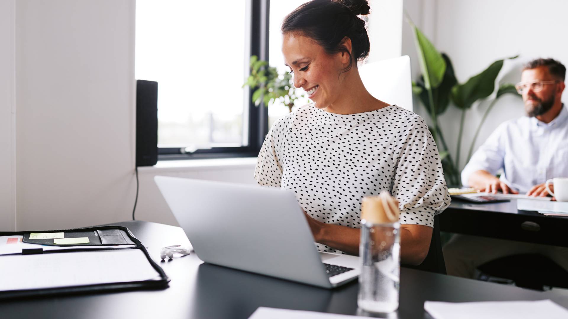 A woman who is smiling, sits at a desk in the office where she works. She's on her laptop and there's a man in the background sitting behind her at a desk on a computer.