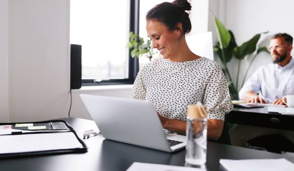 A woman who is smiling, sits at a desk in the office where she works. She's on her laptop and there's a man in the background sitting behind her at a desk on a computer.