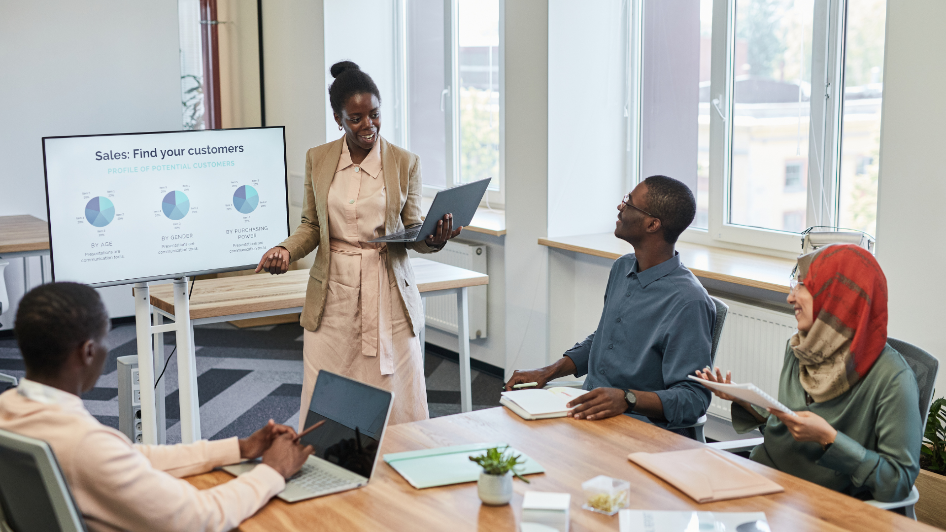 There are four people in a conference room. There is a Black woman who is standing in front of a screen giving a presentation. The other people are seated.