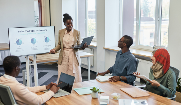There are four people in a conference room. There is a Black woman who is standing in front of a screen giving a presentation. The other people are seated.