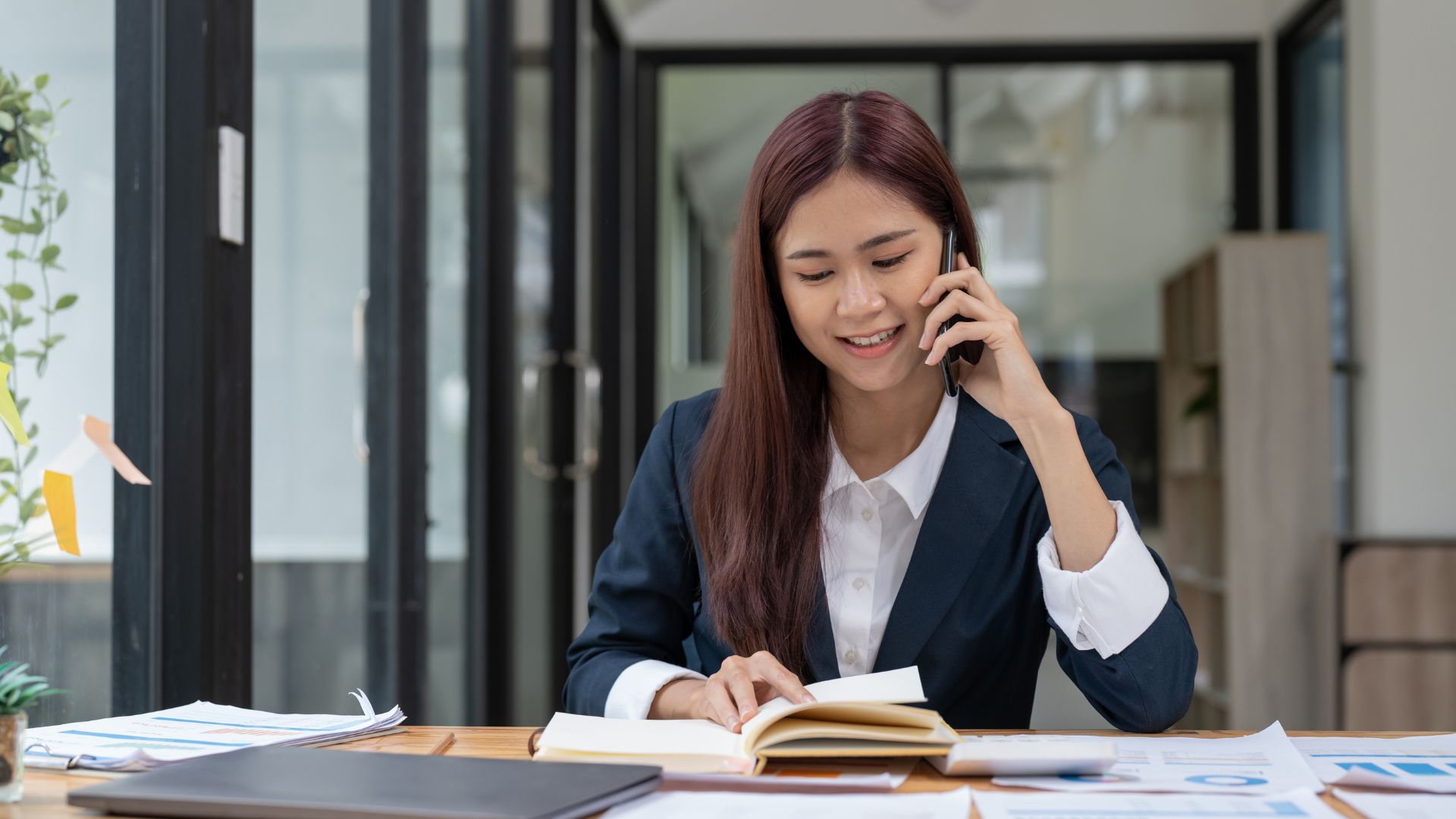 A young woman is talking on her cell phone while looking down at her notebook.
