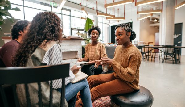 A diverse team of young workers are sitting in an open common area conversing.
