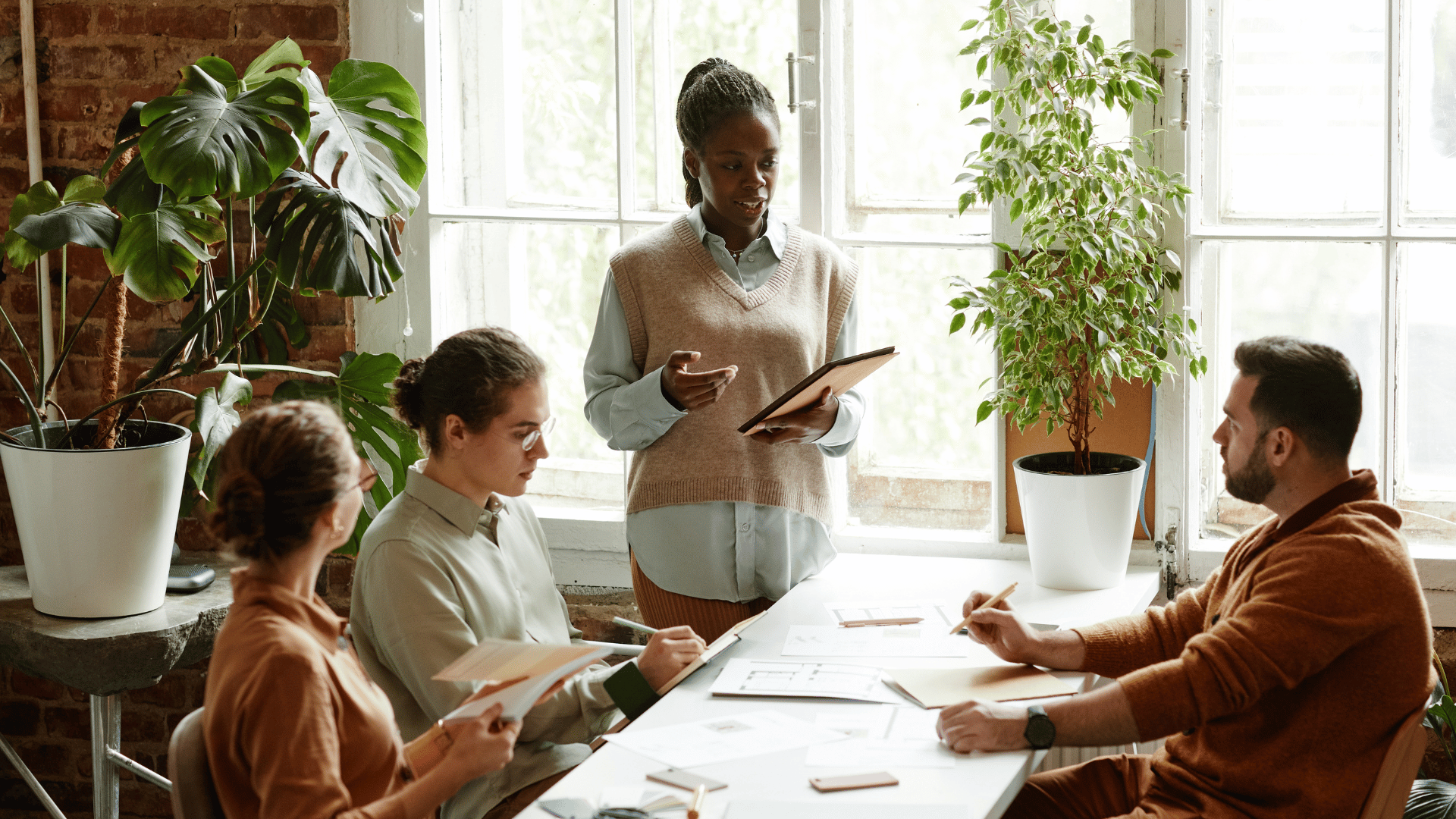 A team of 4 are around a long table. 2 people are sitting and 1 is standing on one side of the table. 1 person is sitting on the other side of the table.