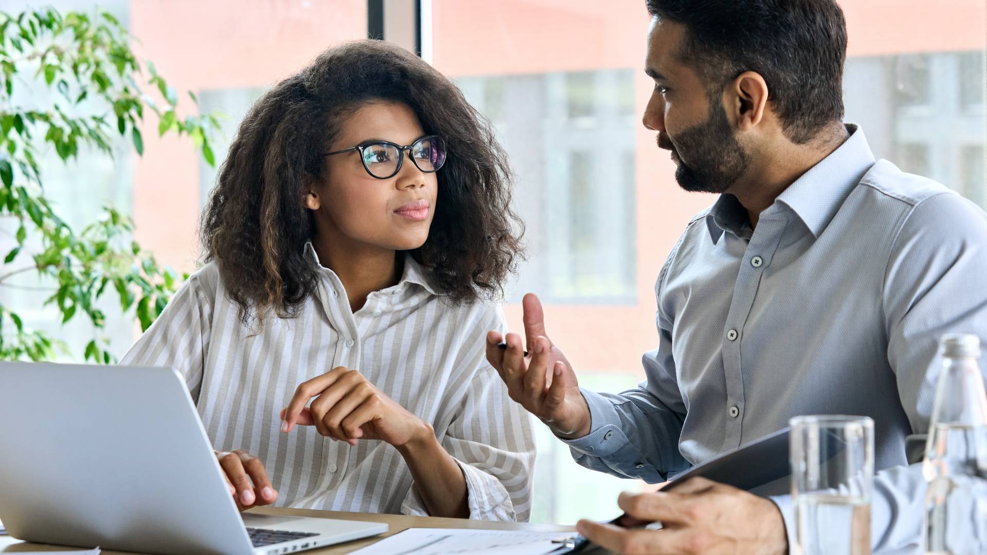 A woman with glasses is listening to a man speak who is speaking with her. She's sitting at a desk with her laptop open.
