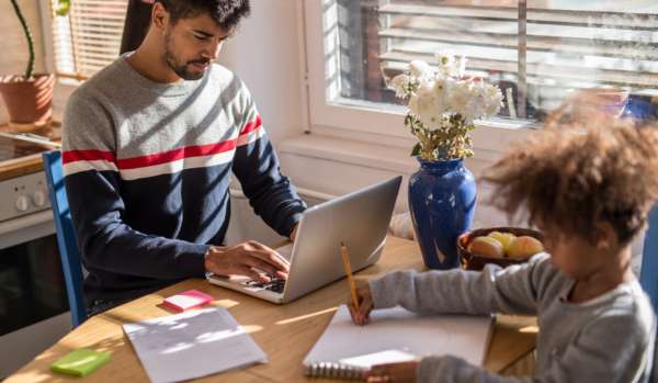 A man works from home at the kitchen table. His child is also at the table, drawing a picture.