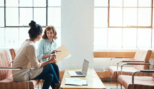 Two women are sitting in an open common area discussing a project. One is holding a folder with papers and the other is holding a coffee.