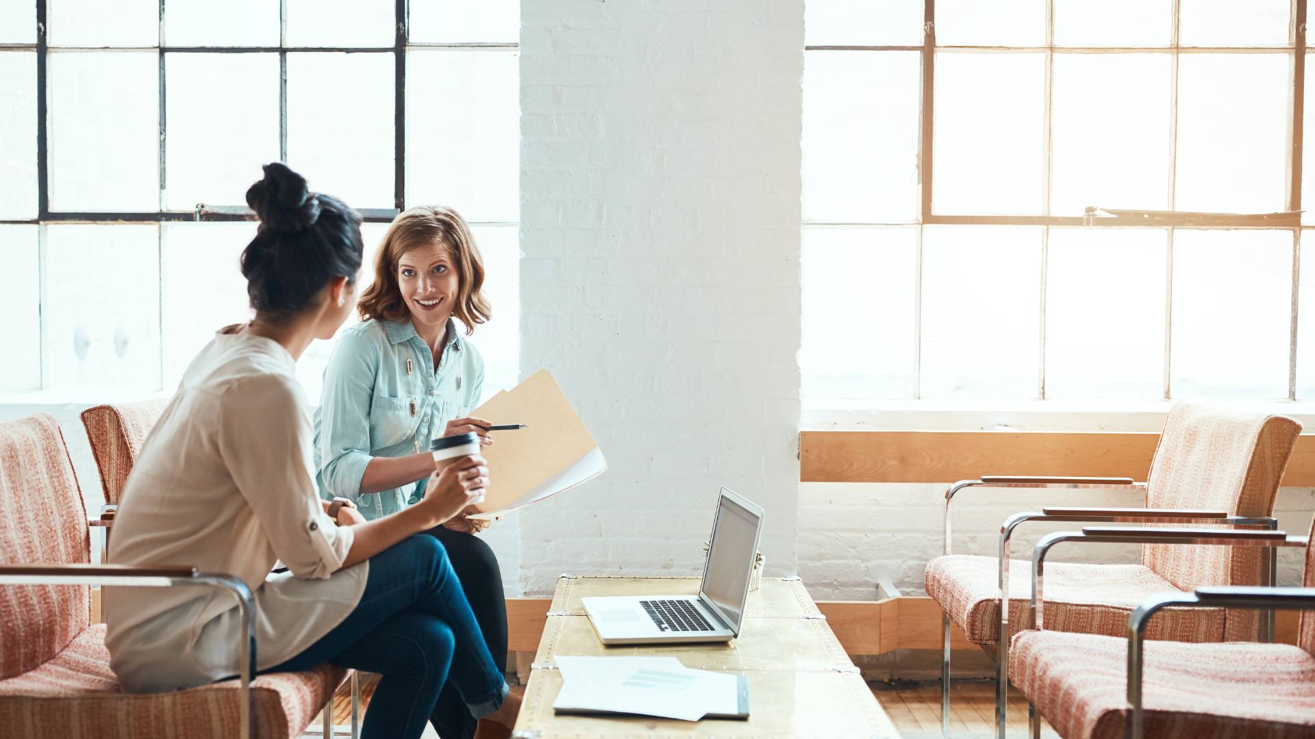Two women are sitting in an open common area discussing a project. One is holding a folder with papers and the other is holding a coffee.