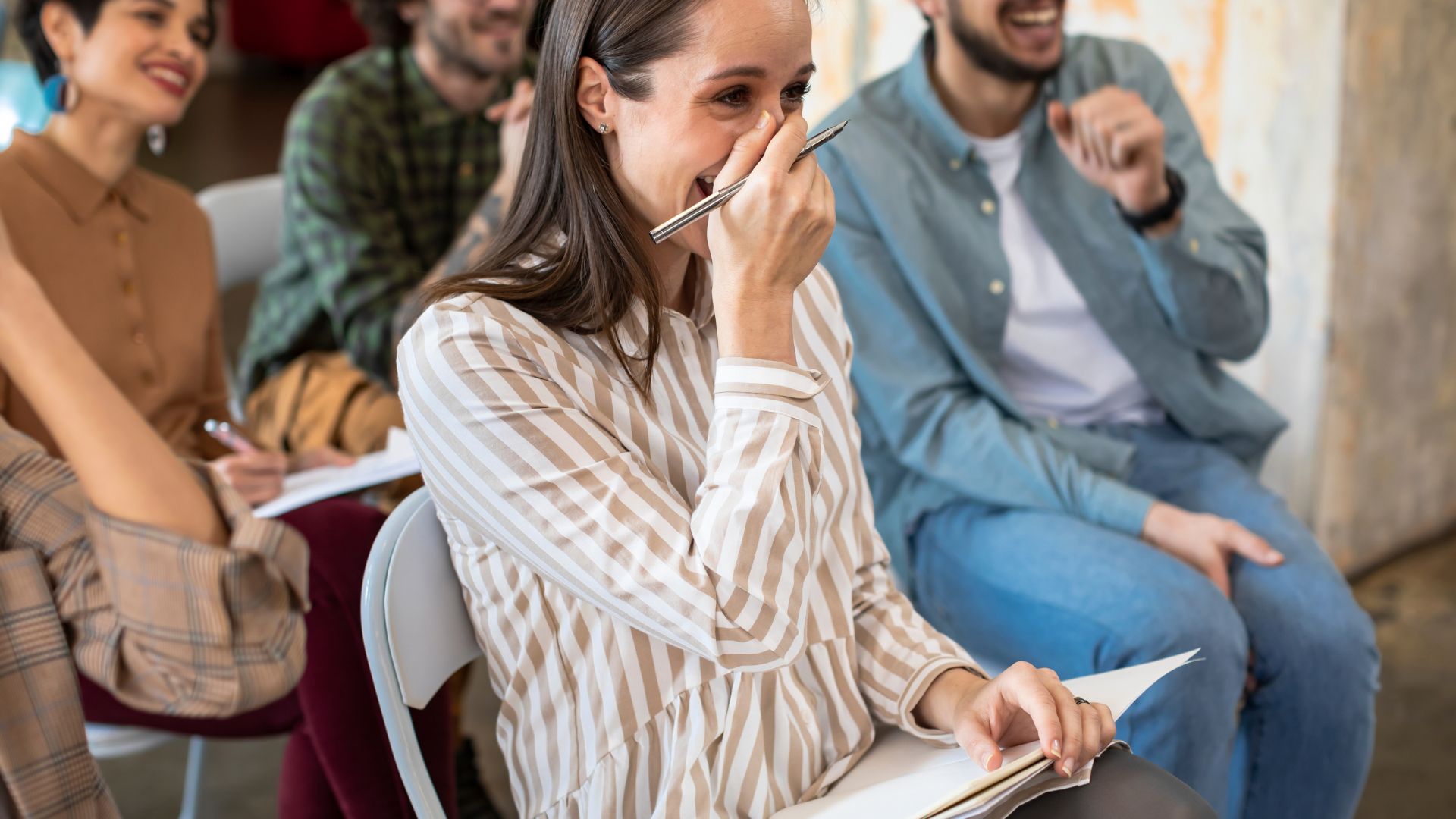 A group of people are sitting in a team building. There is a woman in the forefront with her hand over her mouth, laughing.