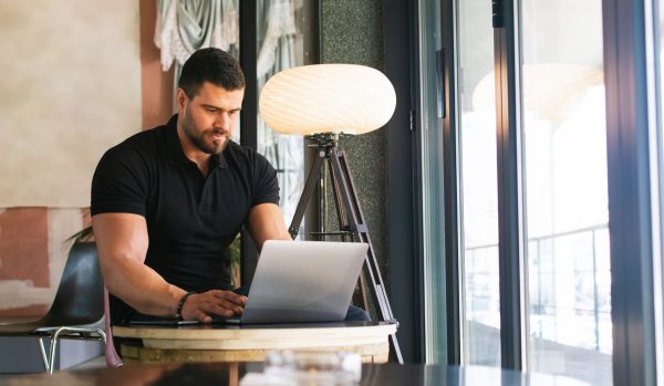 A man is working on his laptop at a small table in a common area near a window.