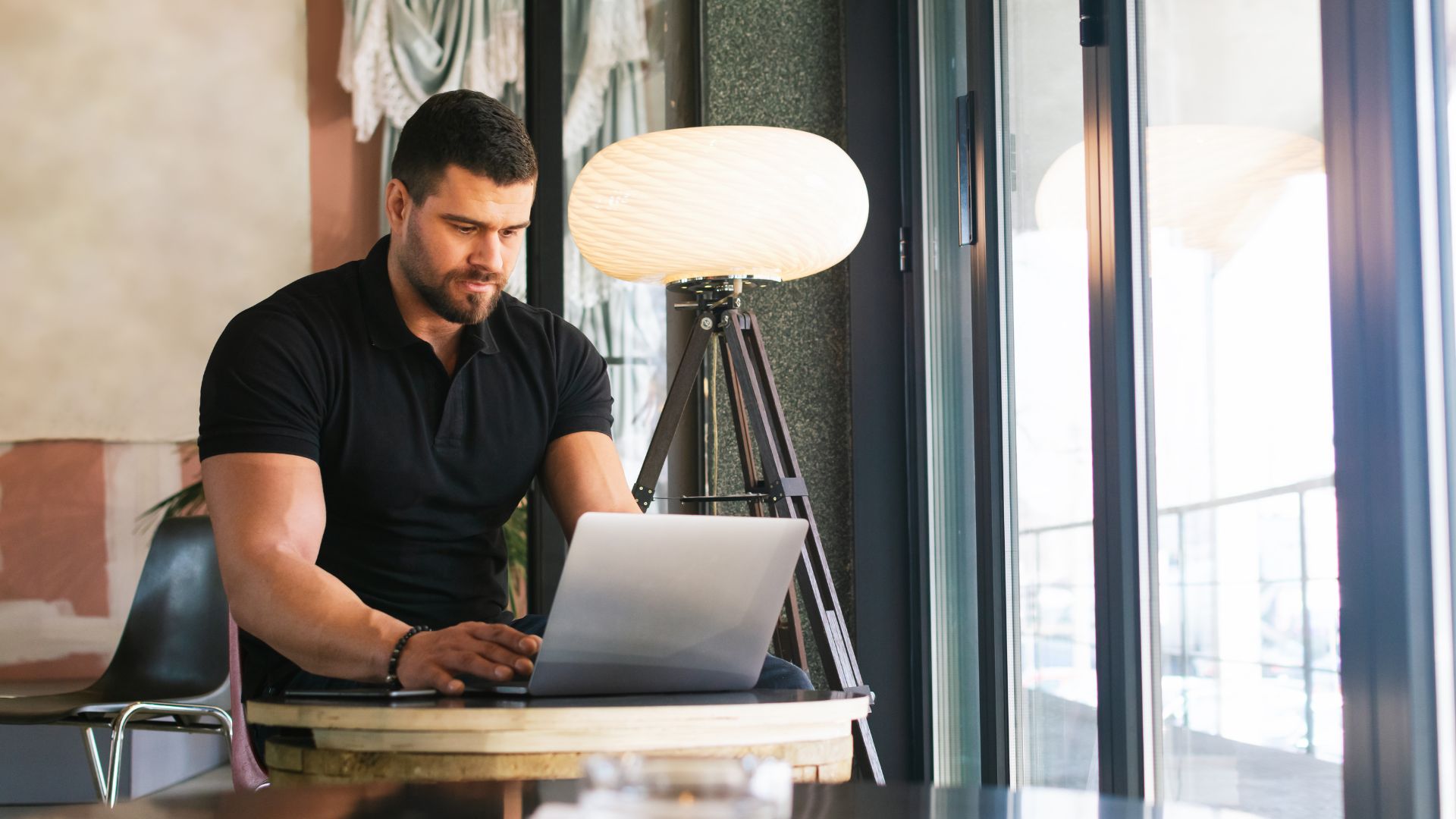 A man is working on his laptop at a small table in a common area near a window.