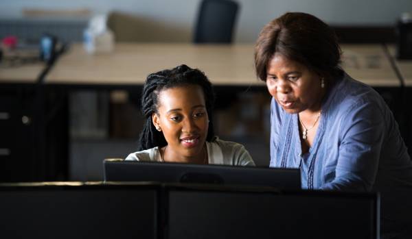 We are looking over a monitor at two woman - one standing one sitting. The older woman is standing and pointing at something on the younger woman's monitor.