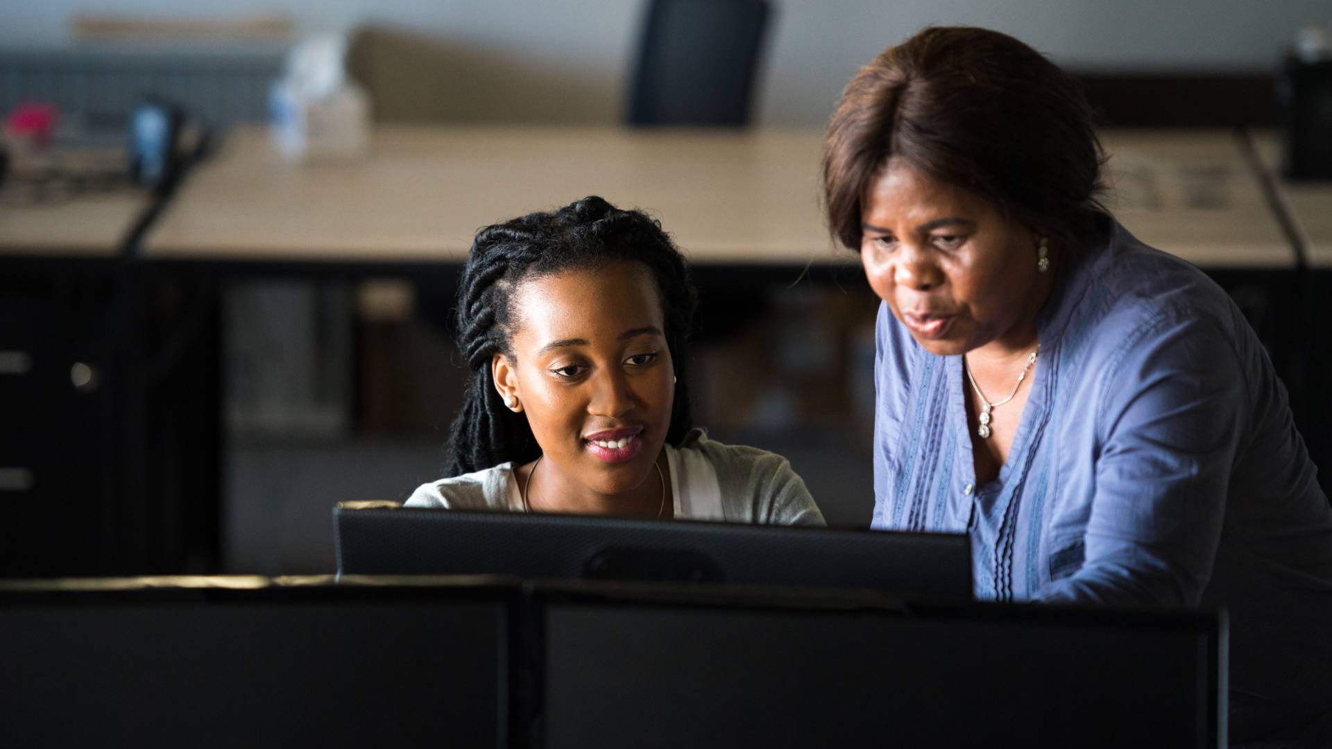 We are looking over a monitor at two woman - one standing one sitting. The older woman is standing and pointing at something on the younger woman's monitor.