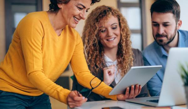 Two women and a man are smiling while looking at a tablet. The woman holding a tablet is standing next to the other two, leaning over.