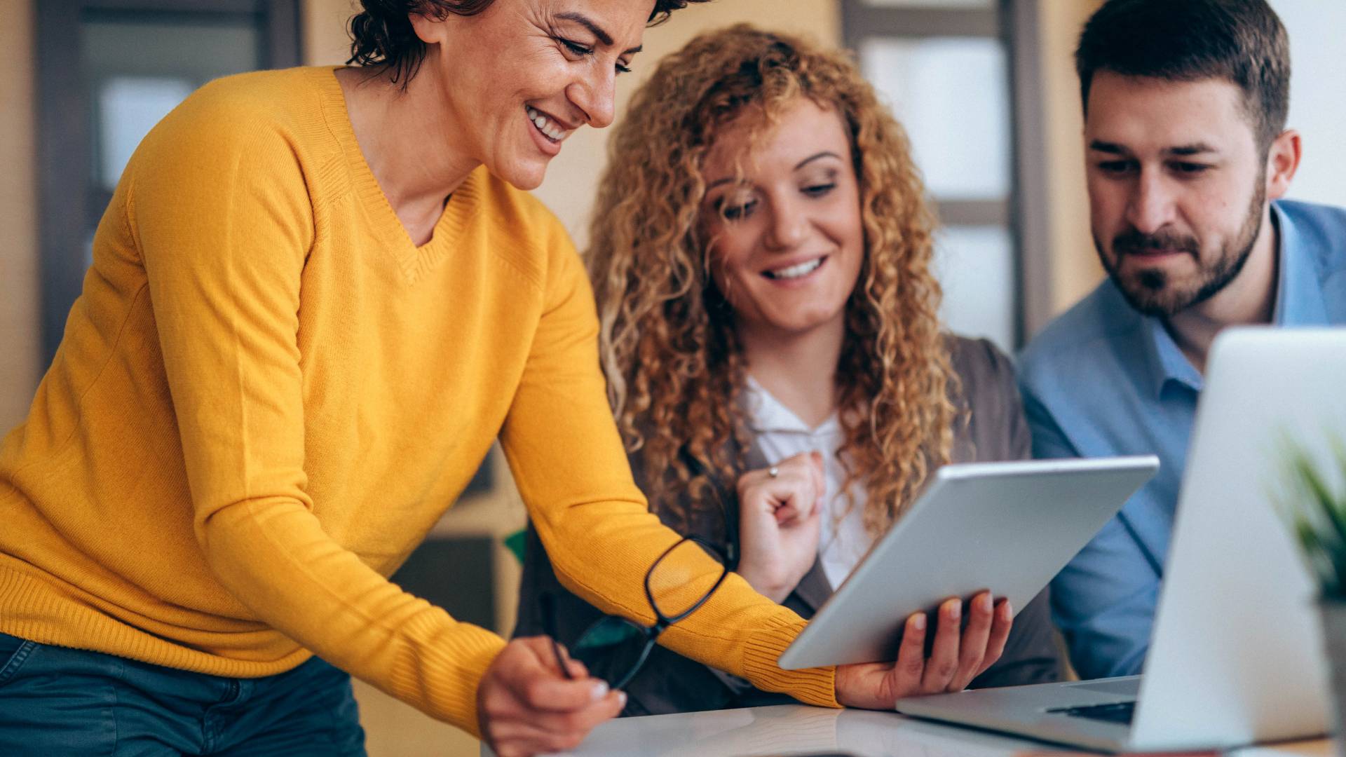Two women and a man are smiling while looking at a tablet. The woman holding a tablet is standing next to the other two, leaning over.