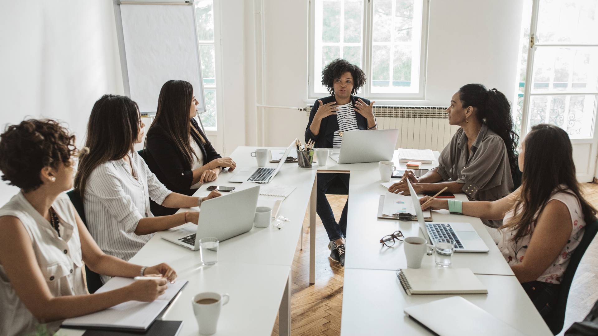 There are a group of six women sitting around a long table engaged in a meeting. There is one woman sitting at the head of the table speaking with the others.