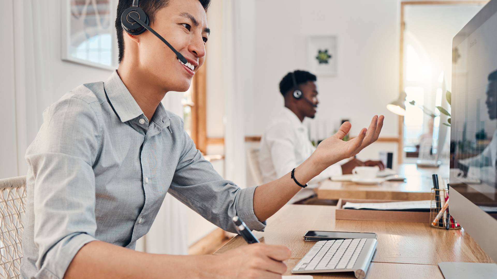 A young salesperson is smiling while talking on his headset at his computer.