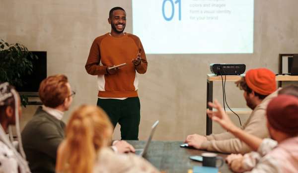 A young man is presenting to a diverse team of people. He is standing, while the team is sitting around a conference team.