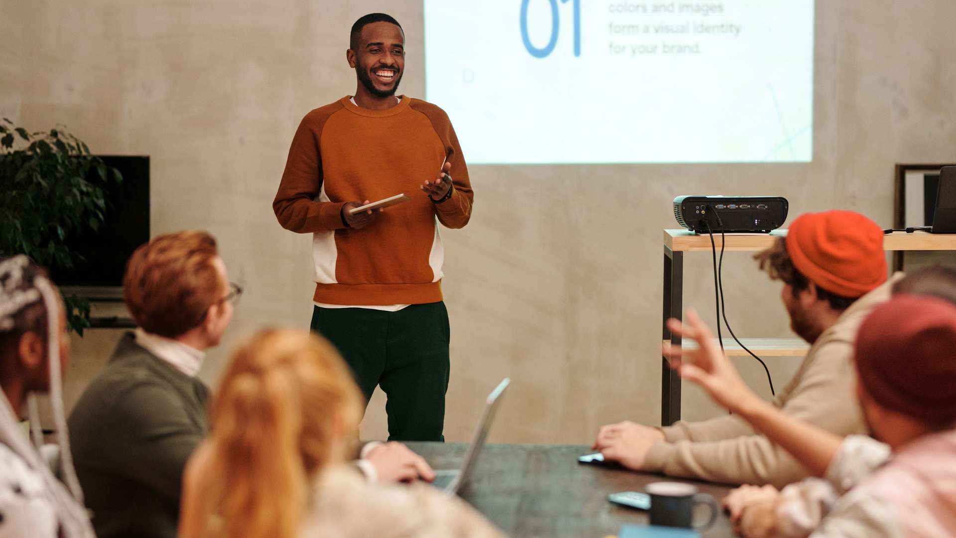 A young man is presenting to a diverse team of people. He is standing, while the team is sitting around a conference team.