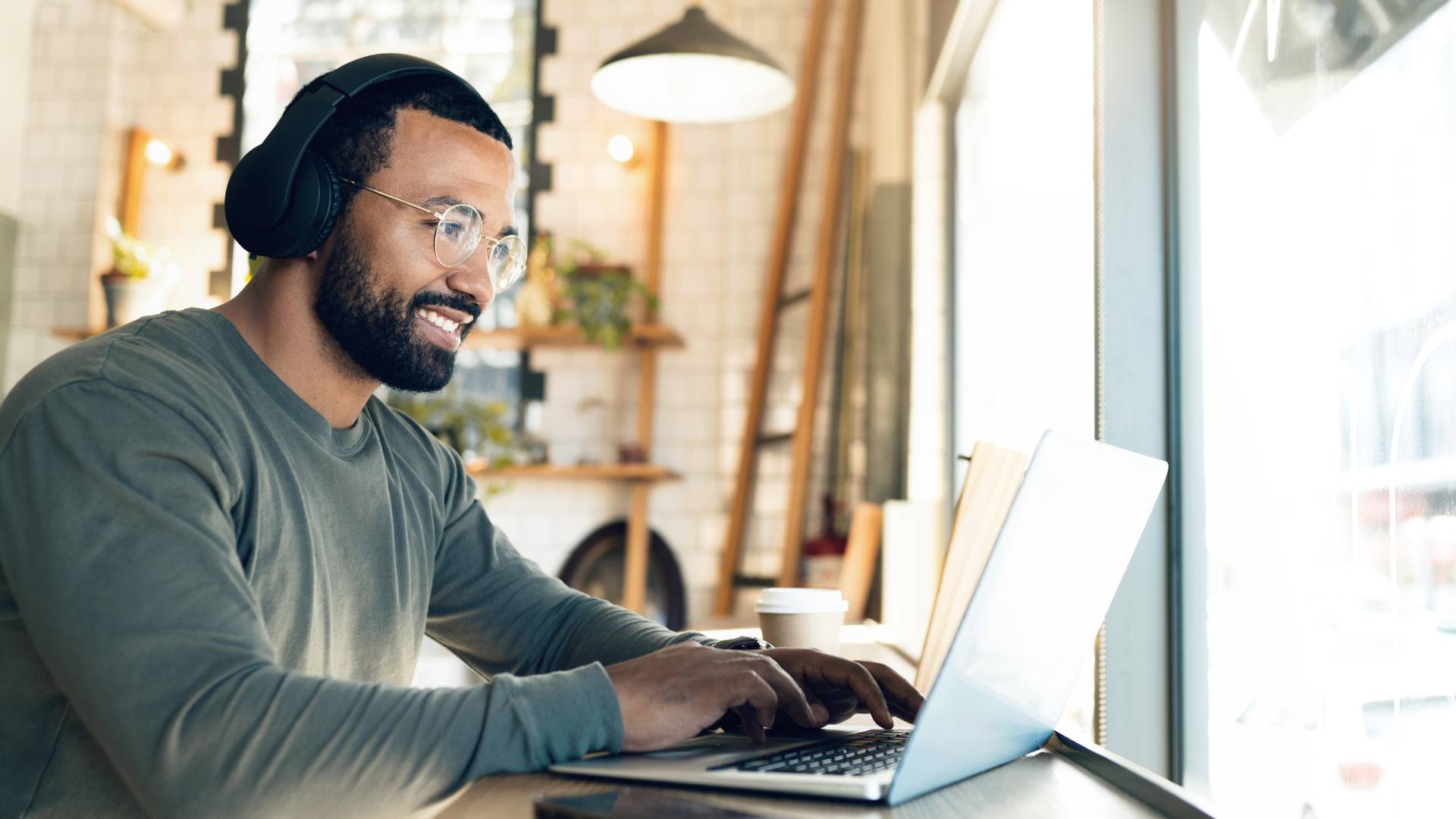 A young man with headphones and glasses is working on his laptop in a coffee shop.