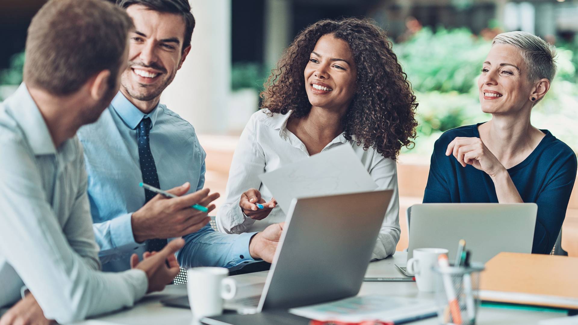 A group of diverse people are laughing and smiling around a laptop on a conference table.